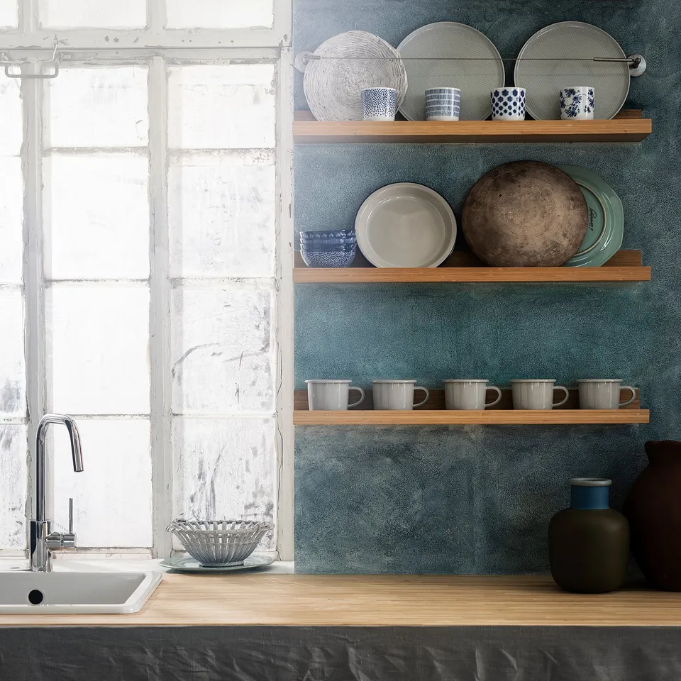 Kitchen counter with sink, wooden shelves holding mugs, plates, and decorative vases against a blue wall.