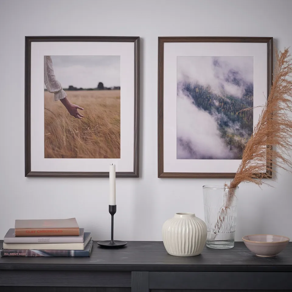 Two framed landscape photos hang on a wall above a dark wooden table with decorative items, including a candle, books, vases, and a bowl.