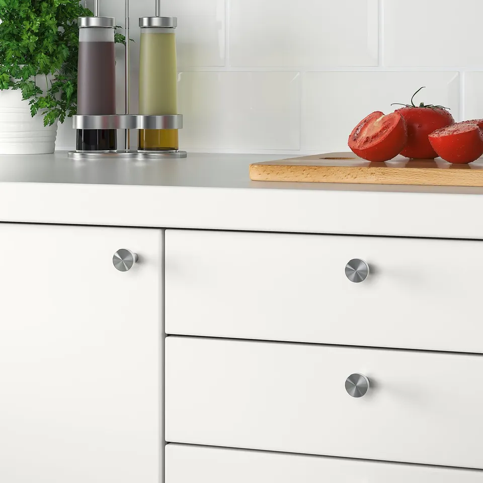 Modern kitchen with white cabinets featuring stainless steel BAGGANÄS knobs. Counter shows tomatoes, herbs, and oil dispensers.