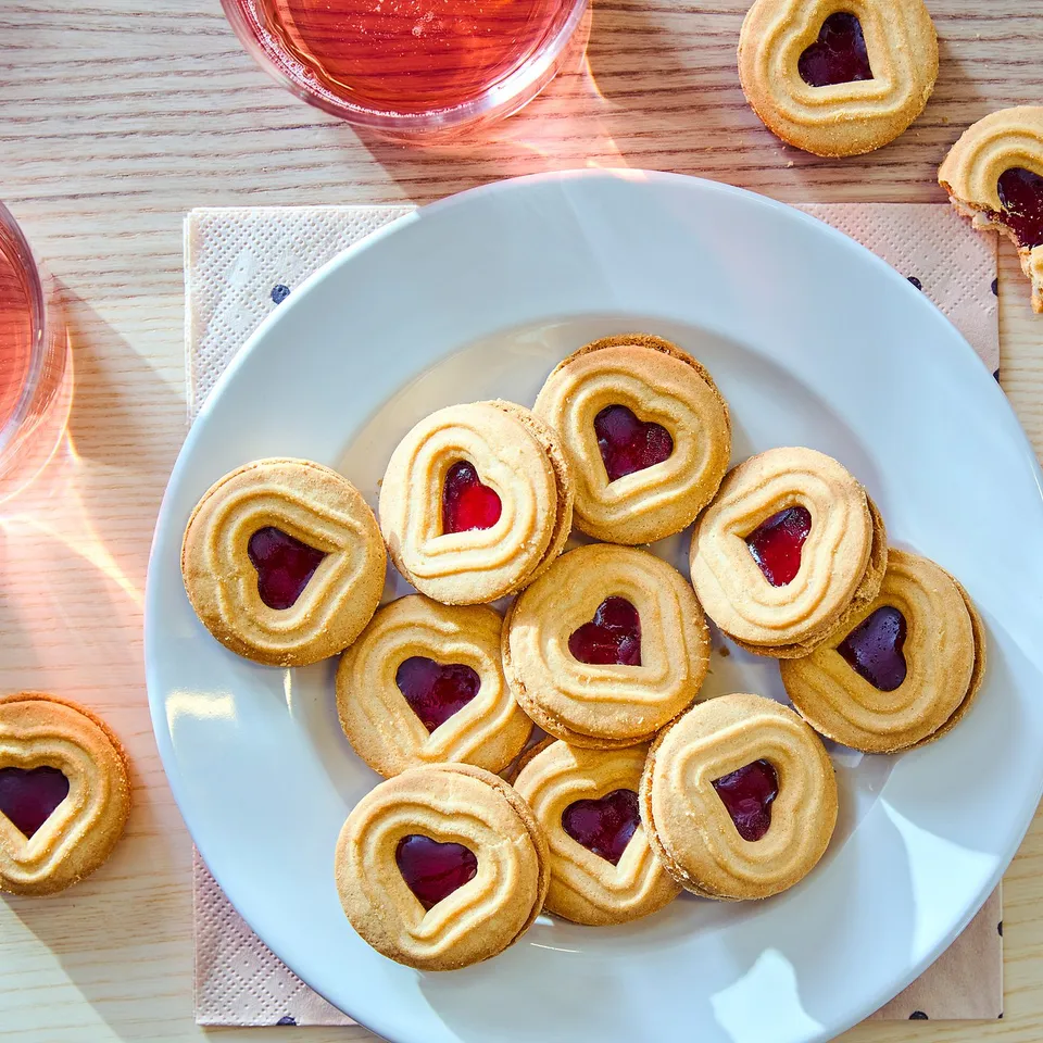 Plate of heart-shaped jam-filled cookies, golden-brown, with pinkish-red jelly centres, and glasses of pink liquid beside it.