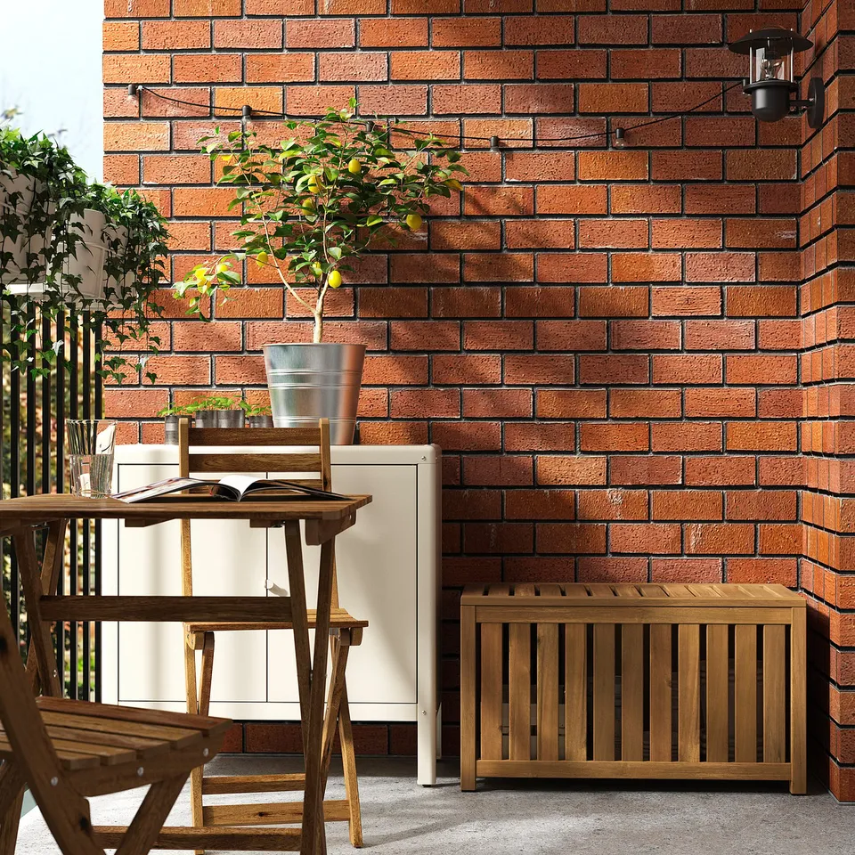 Small wooden outdoor storage box sits against brick wall. Holds items, doubles as extra seat. Perfect for balconies.