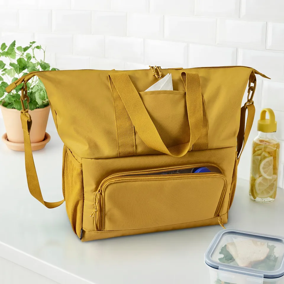 Large mustard-yellow KLOTPIL bag on counter with plant, drink bottle, and lunch box. Has multiple compartments and adjustable straps.
