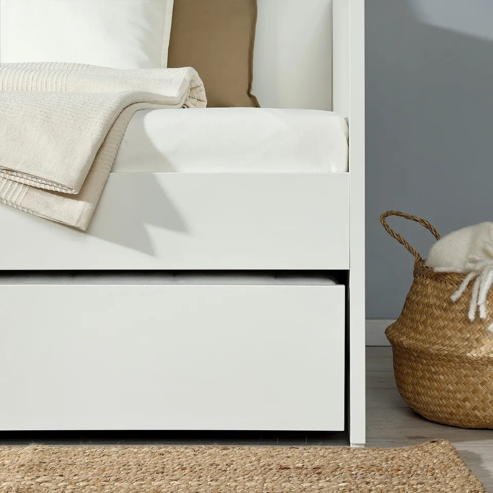 White trundle bed system with a white woven basket beside it. Daybed with a foldable trundle for extra sleep space, shown in a minimalist room.