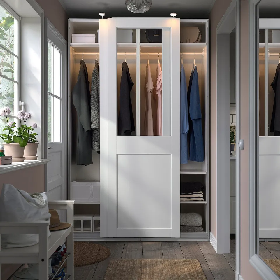 Modern white PAX wardrobe with glass panels, sliding doors, and integrated lighting, efficiently organising clothes in a bedroom.