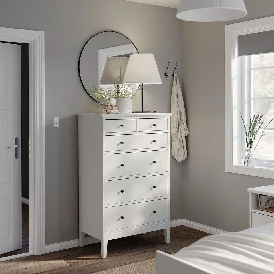 Modern bedroom IDANÄS white dresser with six drawers, topped with a lamp and mirror. Wooden floor and grey walls complete the serene look.