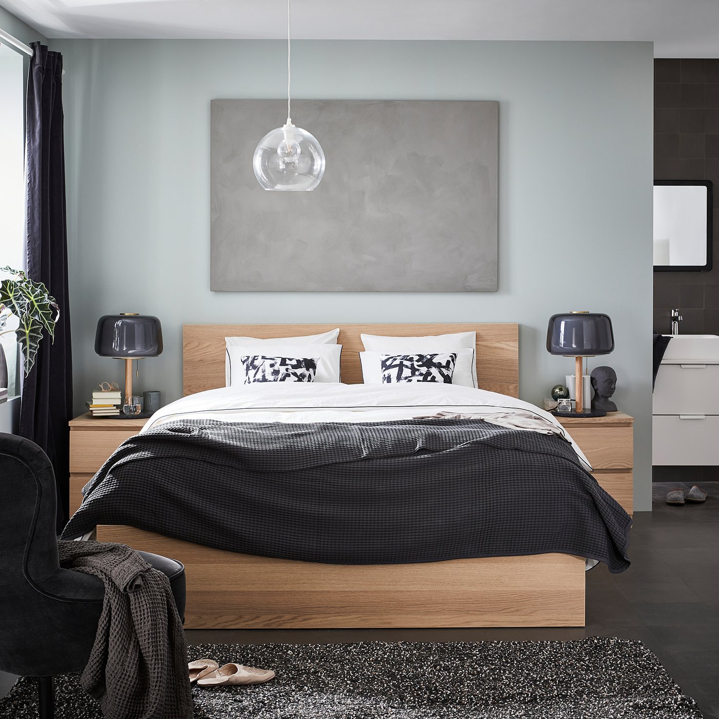 Modern bedroom with blue walls, black decor. Wooden bed with black accents, abstract pillows, pendant lamp, bedside table lamps, and minimalist art.