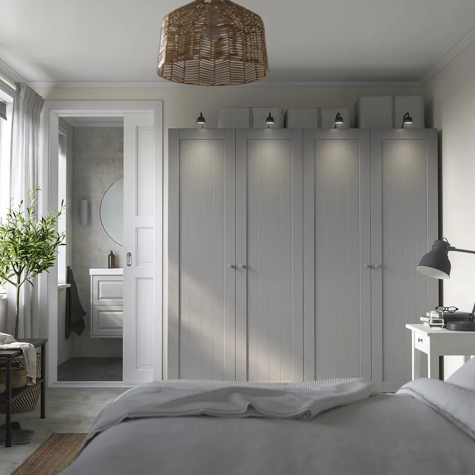 Modern bedroom with large grey PAX wardrobe, woven lampshade, small desk with lamp, and en suite bathroom visible through door.