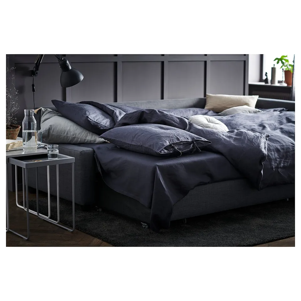 Modern grey sofa-bed with dark bedding, side table, lamp, and plant.
