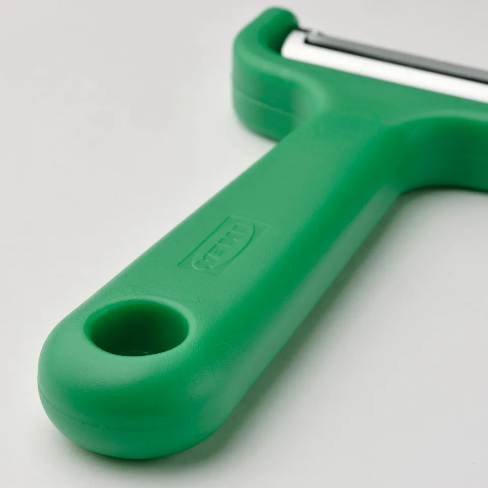 Green plastic-handled peeler with stainless steel blade, resting on a white surface. Ergonomic design with hanging hole in handle.