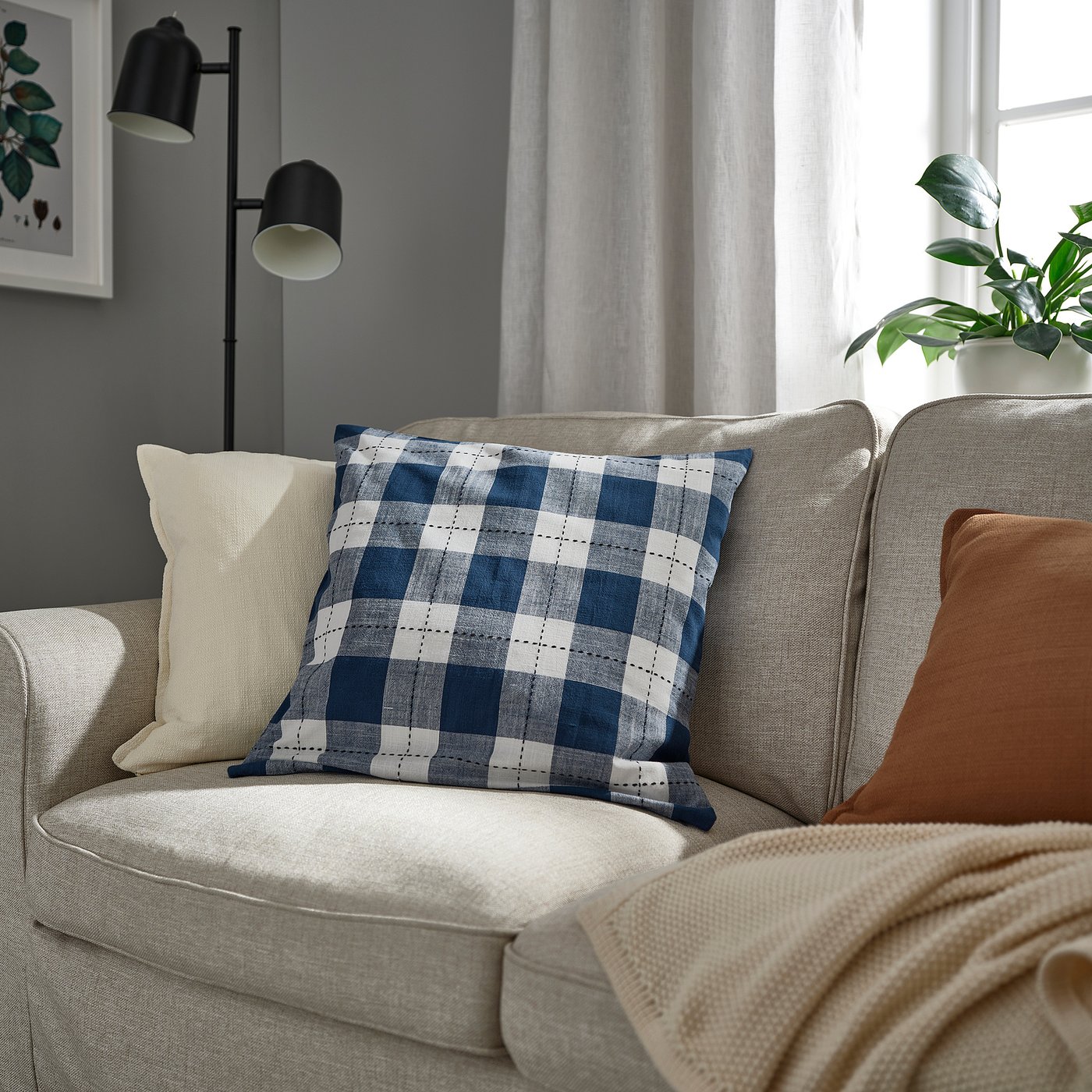 Grey sofa with blue chequered cushion, pillows, blanket, lamp, plant.