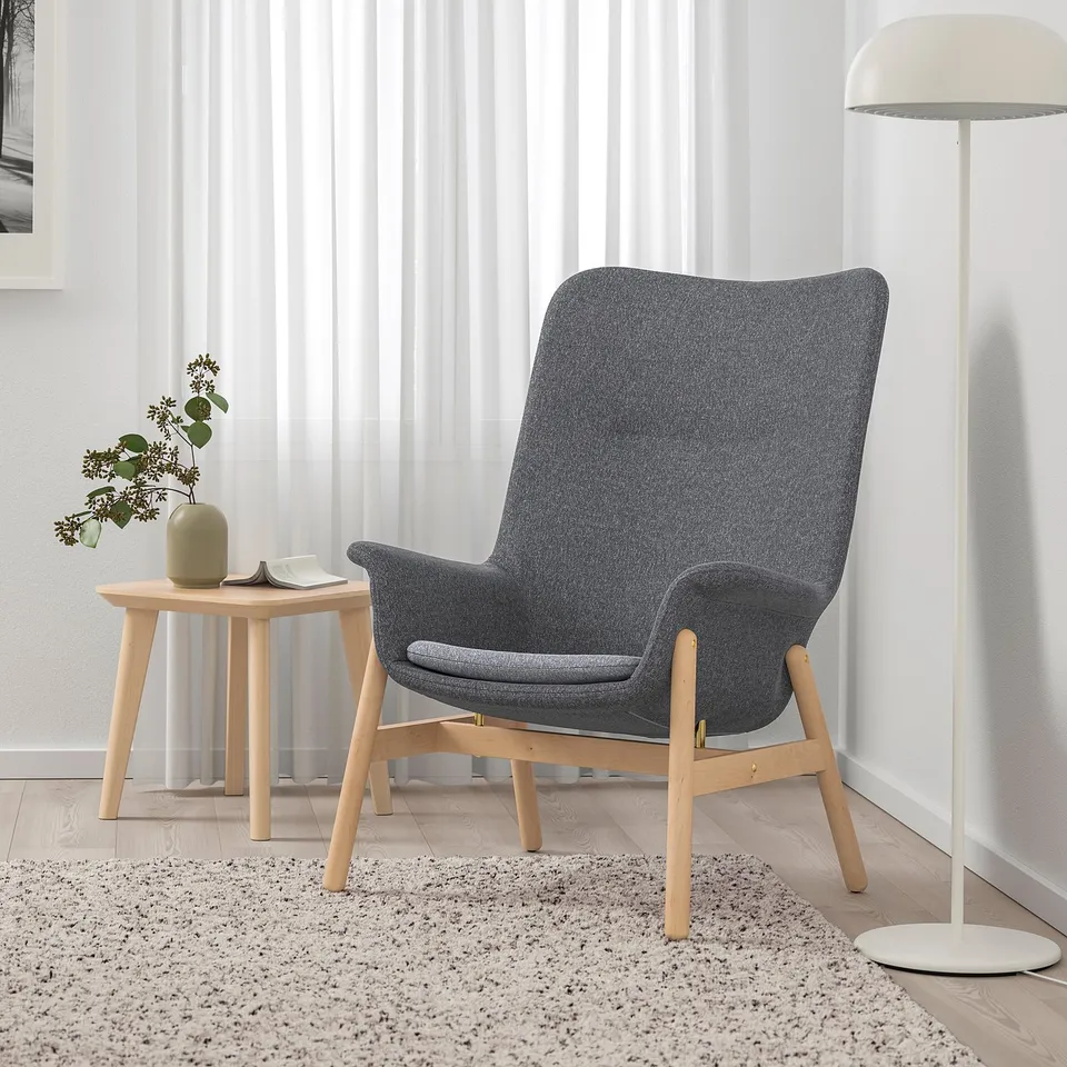 Grey armchair with soft lines, wooden legs, side table with book and lamp.