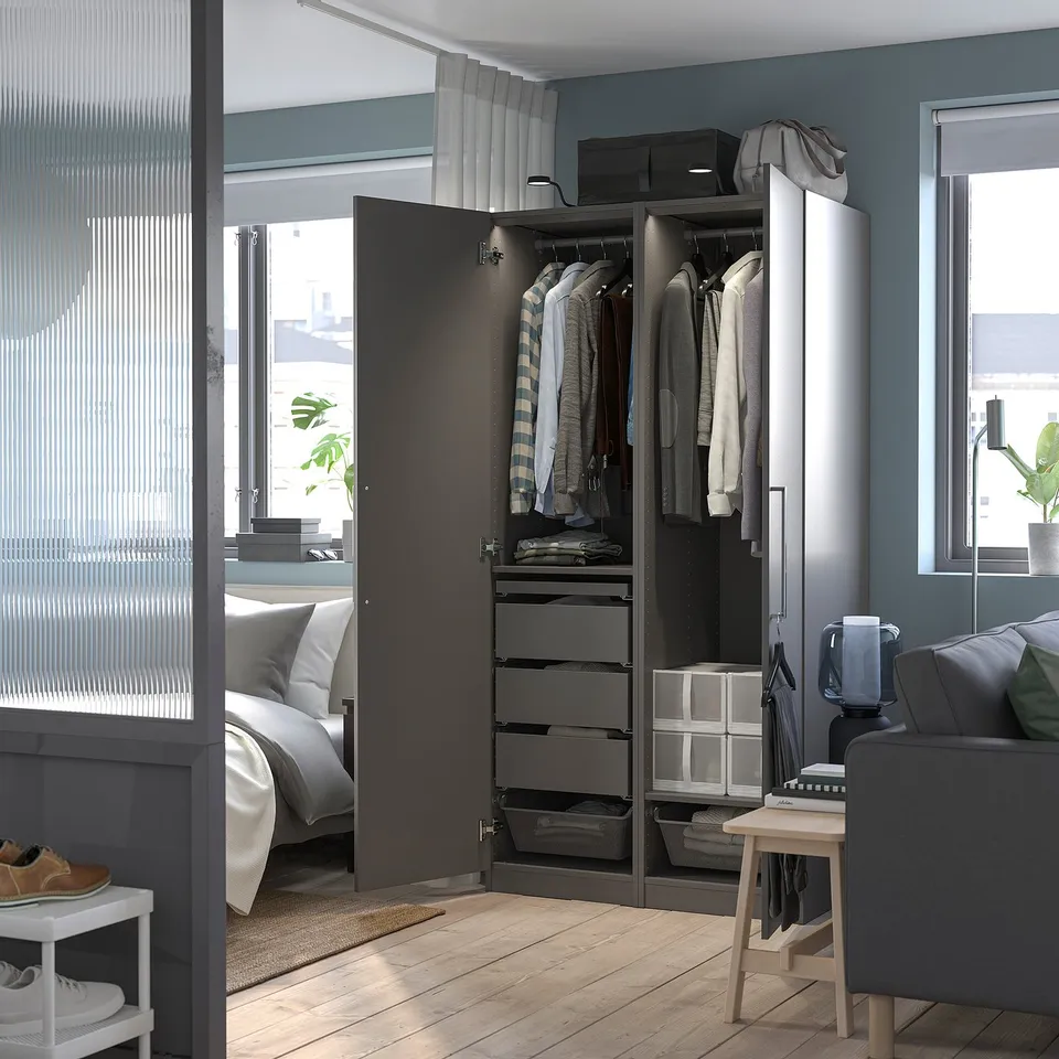 Modern bedroom with open grey wardrobe showing clothes and plants near bed.