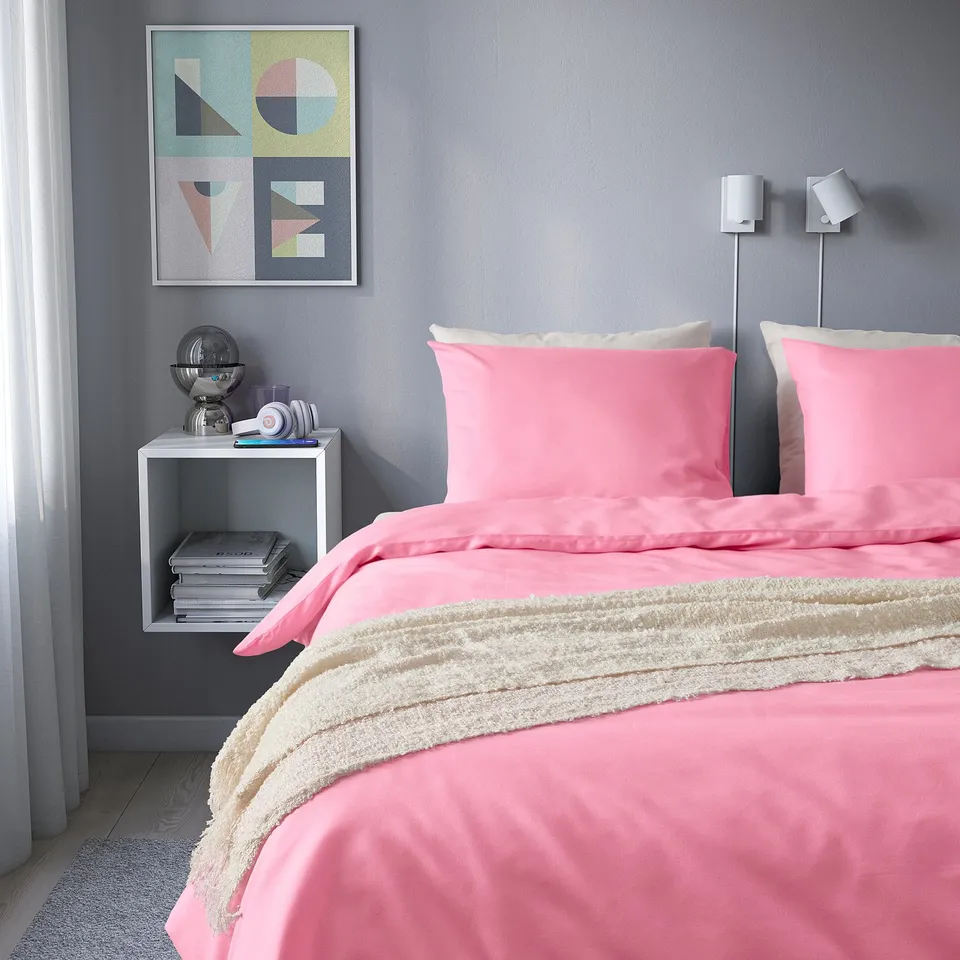 Cosy bedroom with PILTANDVINGE pink duvet & cream pillows, grey wall art, side table with books, lamp, and globe.