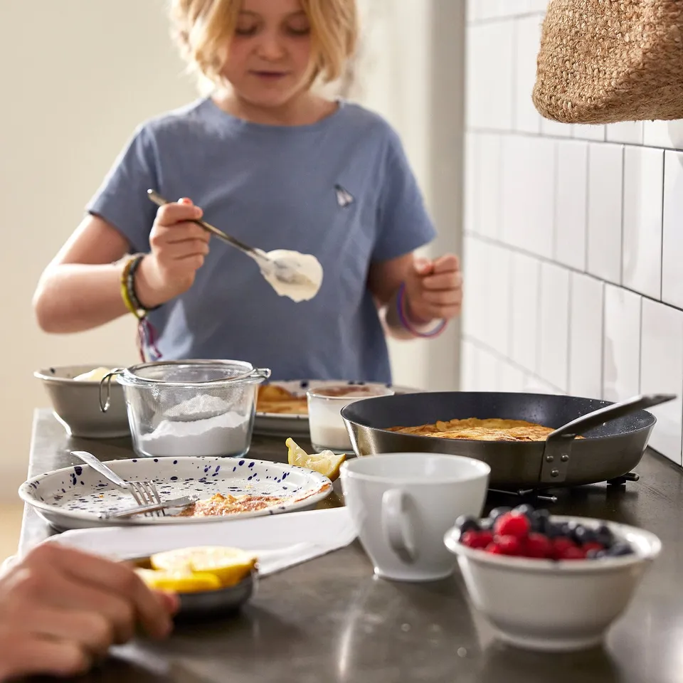 A young girl cooks pancakes in a non-stick frying pan on a stove, surrounded by ingredients and serving dishes.