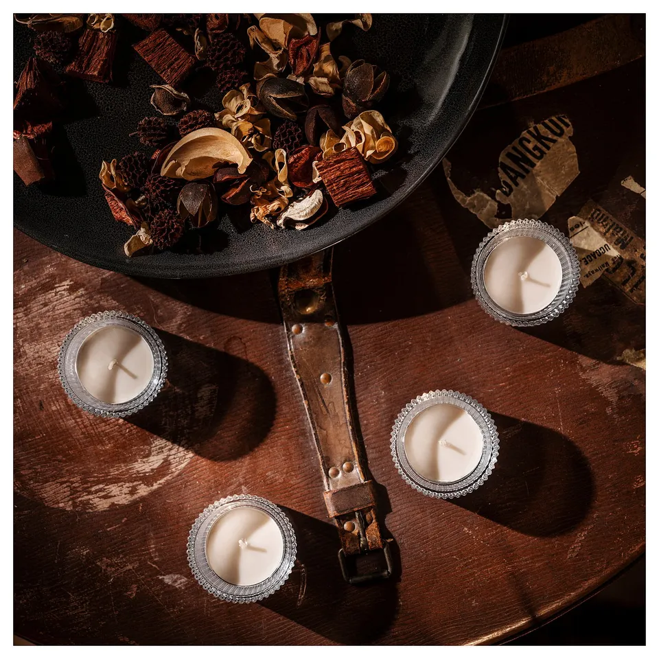 Four candles on wooden table with dried flowers and pinecones in background, illuminated by soft light.