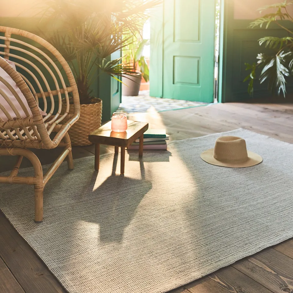 A cosy, sunlit patio with a TIPHEDE blue-gray cotton rug, a wicker chair, a hat, and a candle on a small table, leading to a green door.
