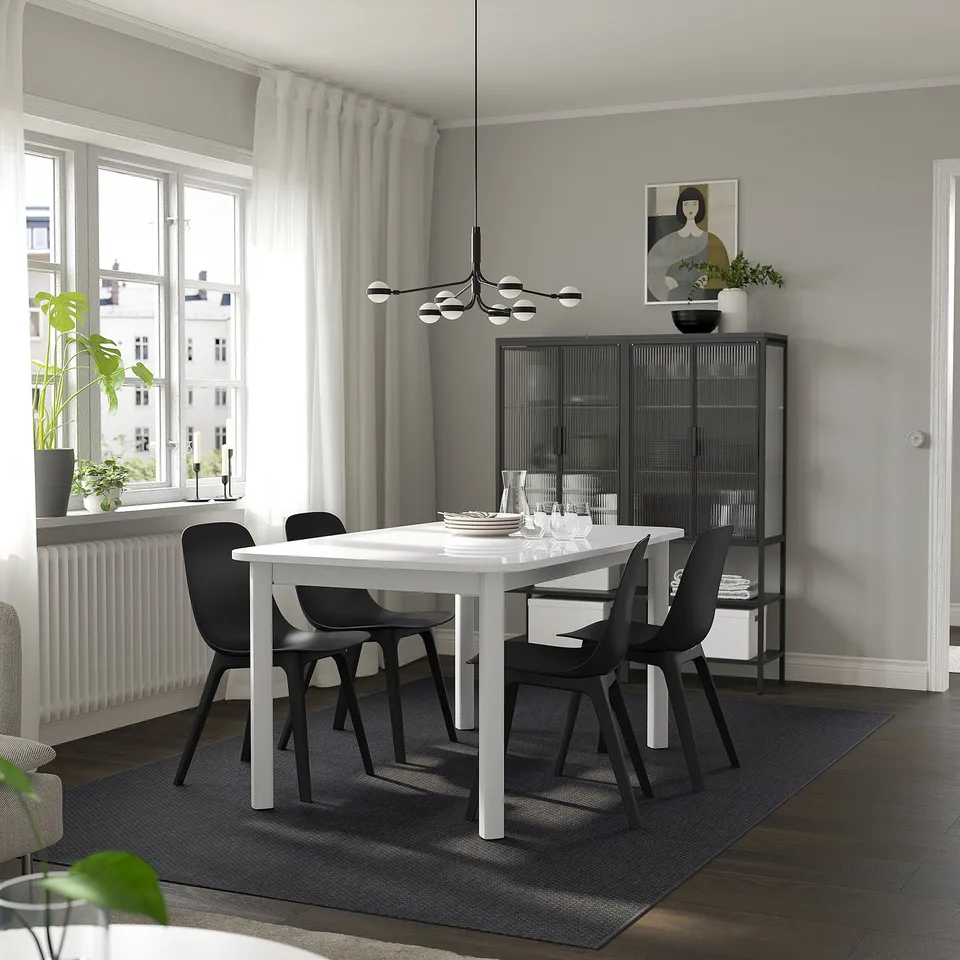 Modern dining room with STRANDTORP table, black chairs, rug, glass cabinet, and plants. Large windows let in natural light.