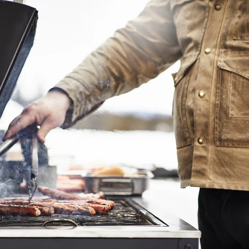 Person grilling sausages on a GRILLSKÄR charcoal grill with black lid and stainless steel grid. Outdoor setting.