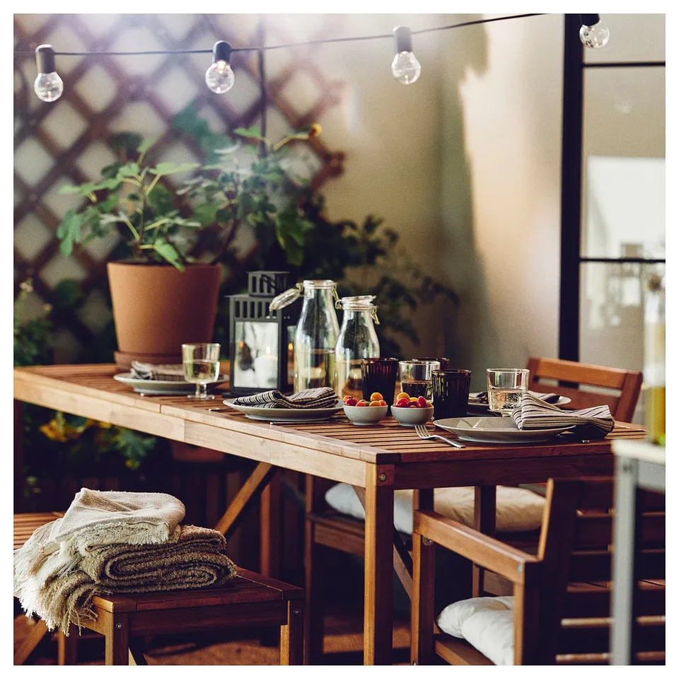 Outdoor table set with dinnerware, lights, plant, and blankets.