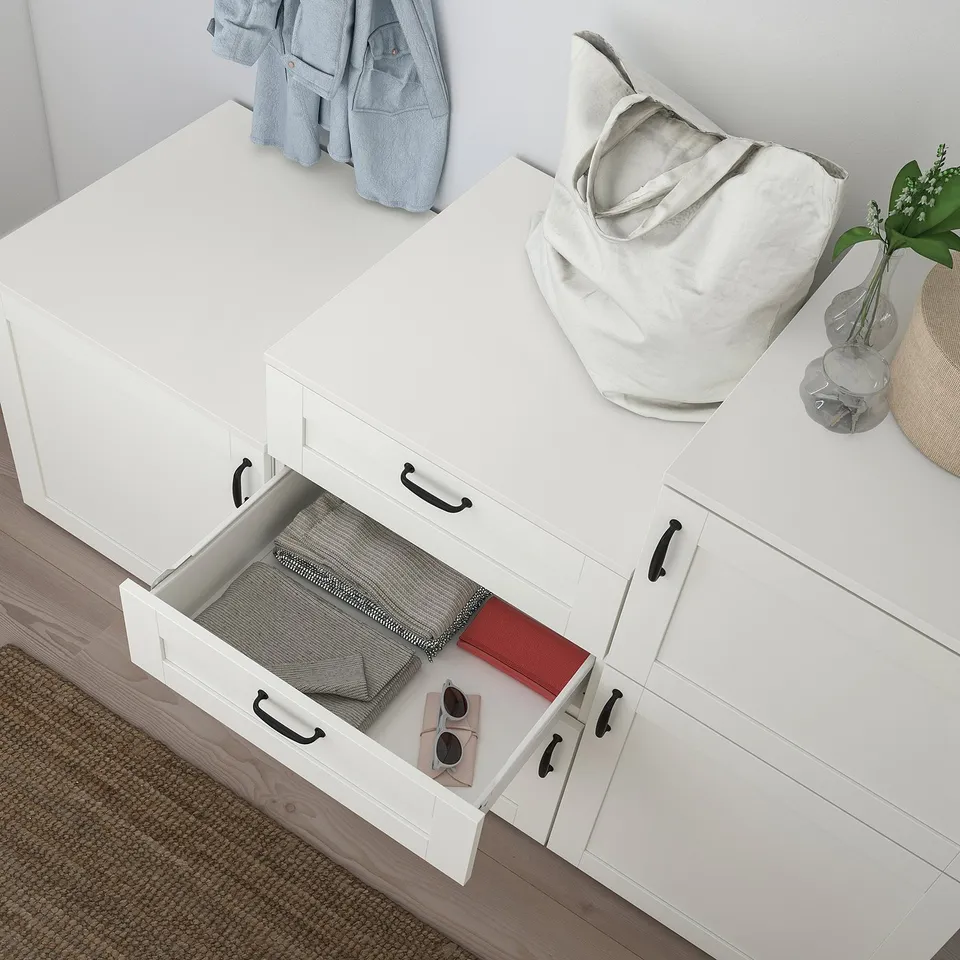 A modern white PLATSA storage unit with a drawer pulled out, displaying clothes, sunglasses, and accessories. The top holds bags and a plant.