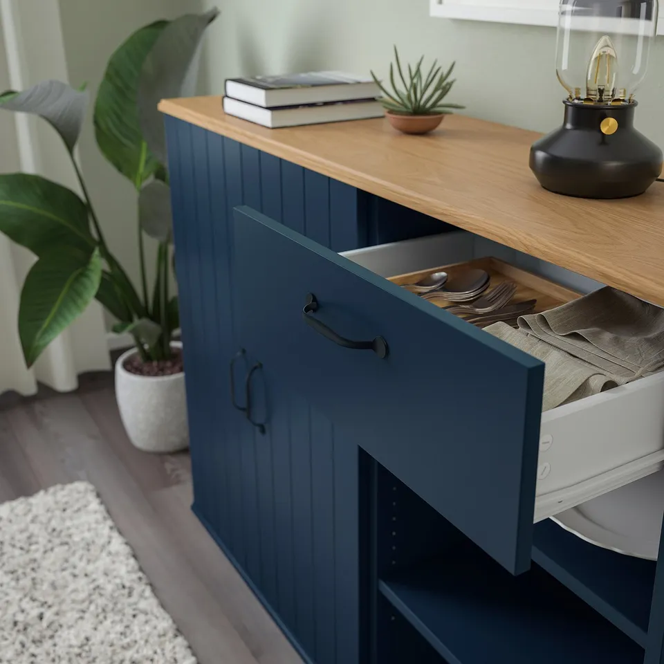 Blue cabinet with open drawer revealing cutlery and cloths, topped with a wooden surface holding books, a small plant, and a lamp.
