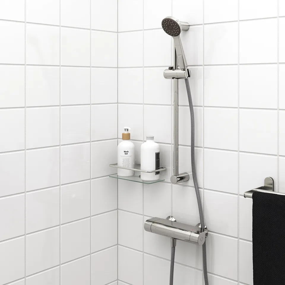 Wall-mounted VALLAMOSSE handshower with chrome, grey hose, and adjustable bracket. Includes glass shelf with products on tiled wall.