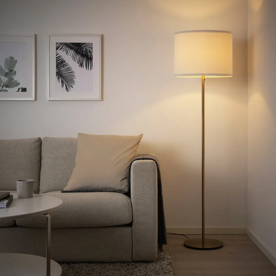Modern living room with grey sofa, large floor lamp SKAFTET in brass tone beside it, emitting warm light. Laundry racks are also visible.