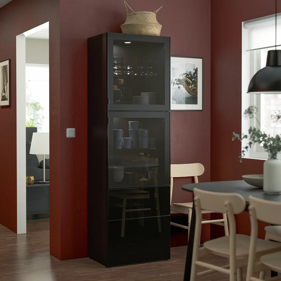 Black BESTÅ storage unit in dining room, taller than nearby chairs, displaying cups and decor, against burgundy wall.