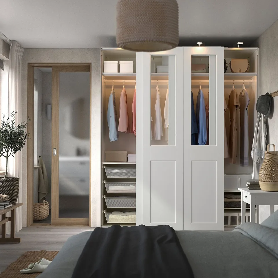 White PAX wardrobe with glass panels and colourful clothes, tall with sleek doors and lighting.