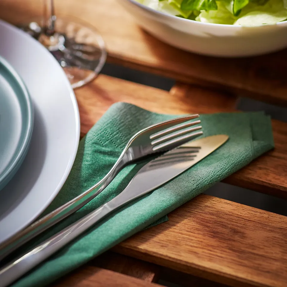Dinner table with white plate, green napkin, fork, knife, and bowl of salad on a wooden table.