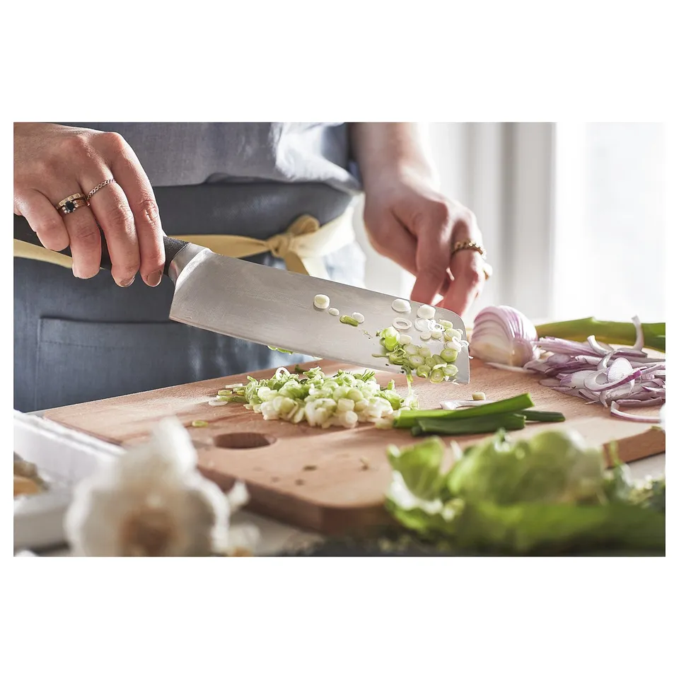 Chopping vegetables on wooden board with knife. Green onions, garlic, red onions visible. Kitchen setting.
