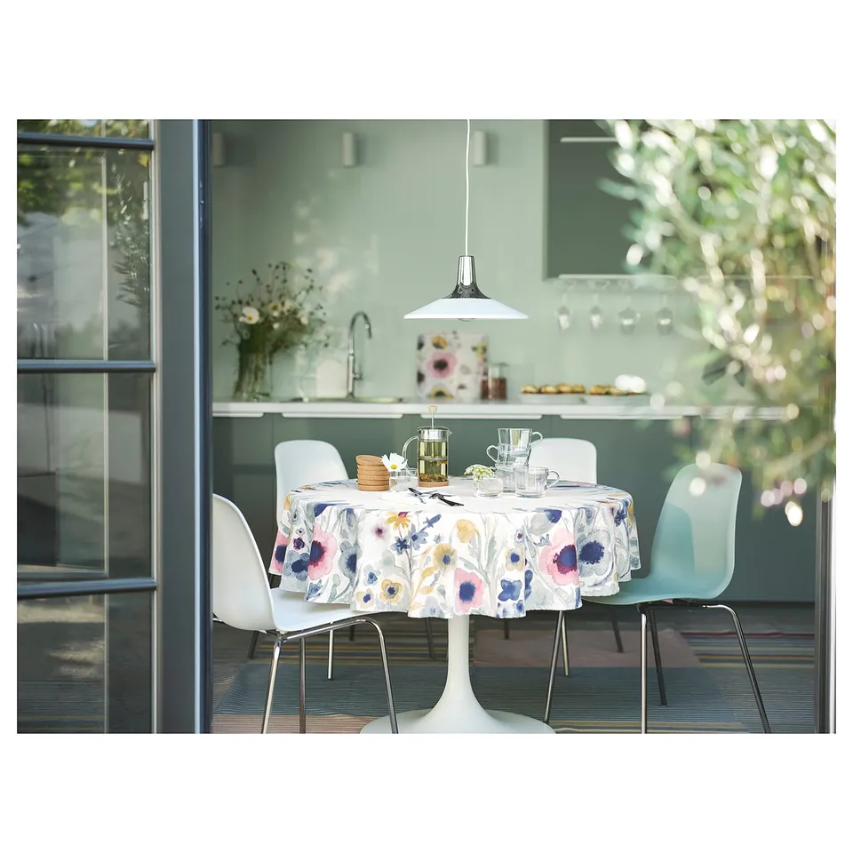 Modern dining scene with white tulip table, colourful floral tablecloth, blue chairs, hanging white lamp, glassware, and green kitchen backsplash.