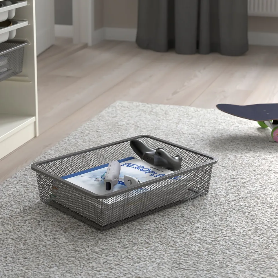 Metal mesh storage box on carpet with gaming controllers and case. TROFAST organisational tool for easy access and tidying.