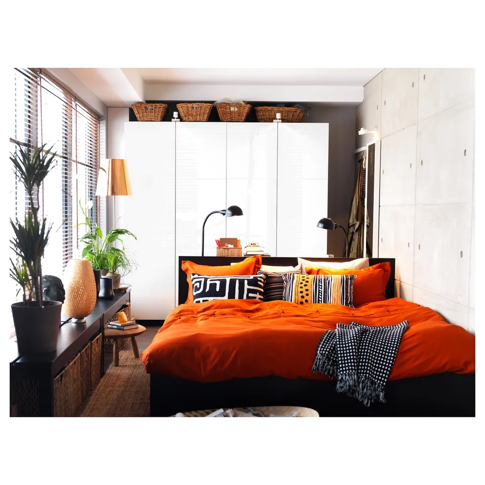 Bright bedroom with orange bedding, patterned pillows, wooden and black furniture, plants, and black lamps.