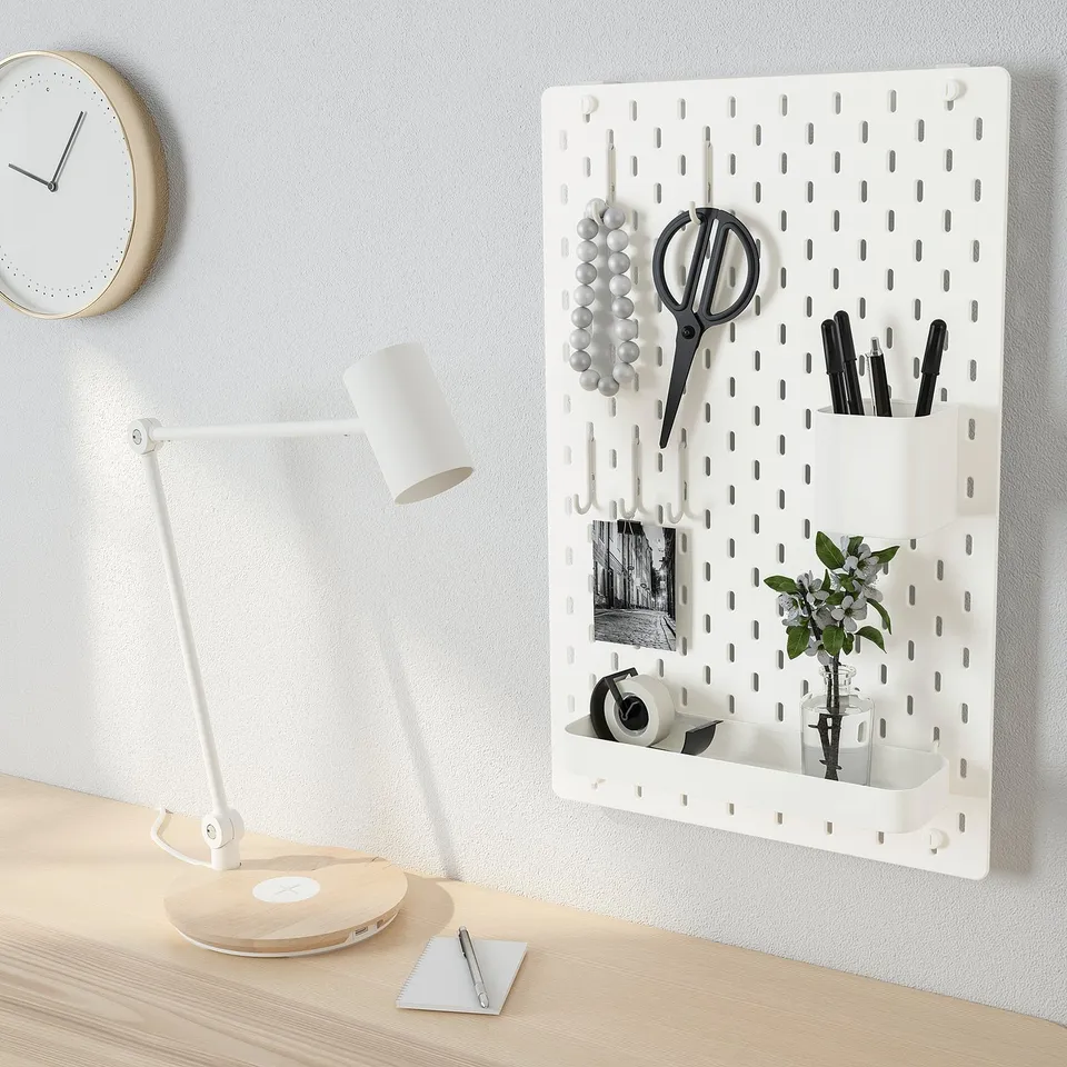 A pegboard organiser with scissors, pens, a plant, and a mirror, alongside a desk with a lamp and a clock on the wall.