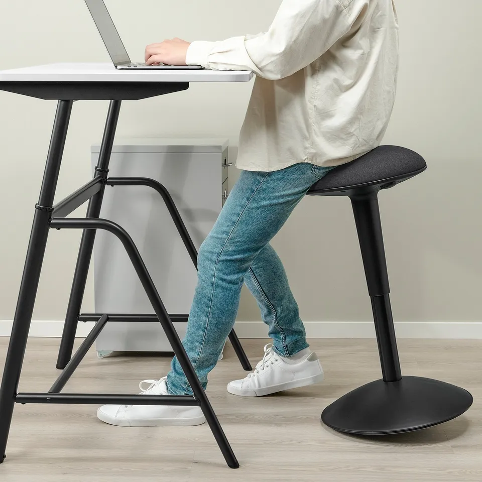 Person sitting on black adjustable stool at desk, typing on laptop.
