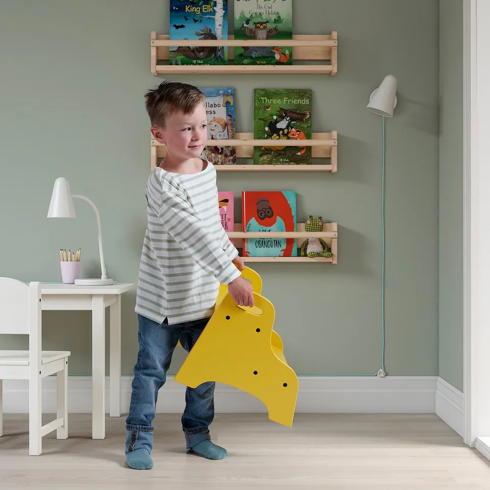 Boy holding yellow TROGEN stool, designed for children. Positioned in a room with bookshelves and a desk.