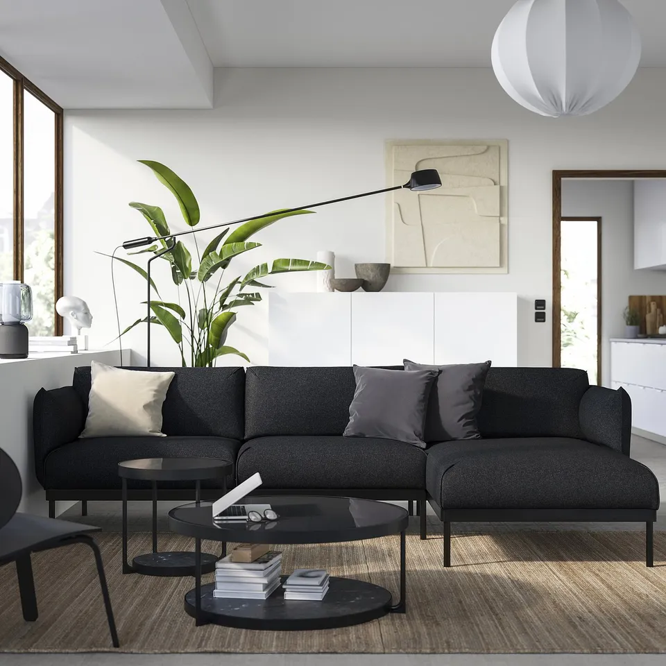 Modern living room with ÄPPLARYD dark grey sofa, black coffee table, large green plant, abstract wall art, and minimalist decor.