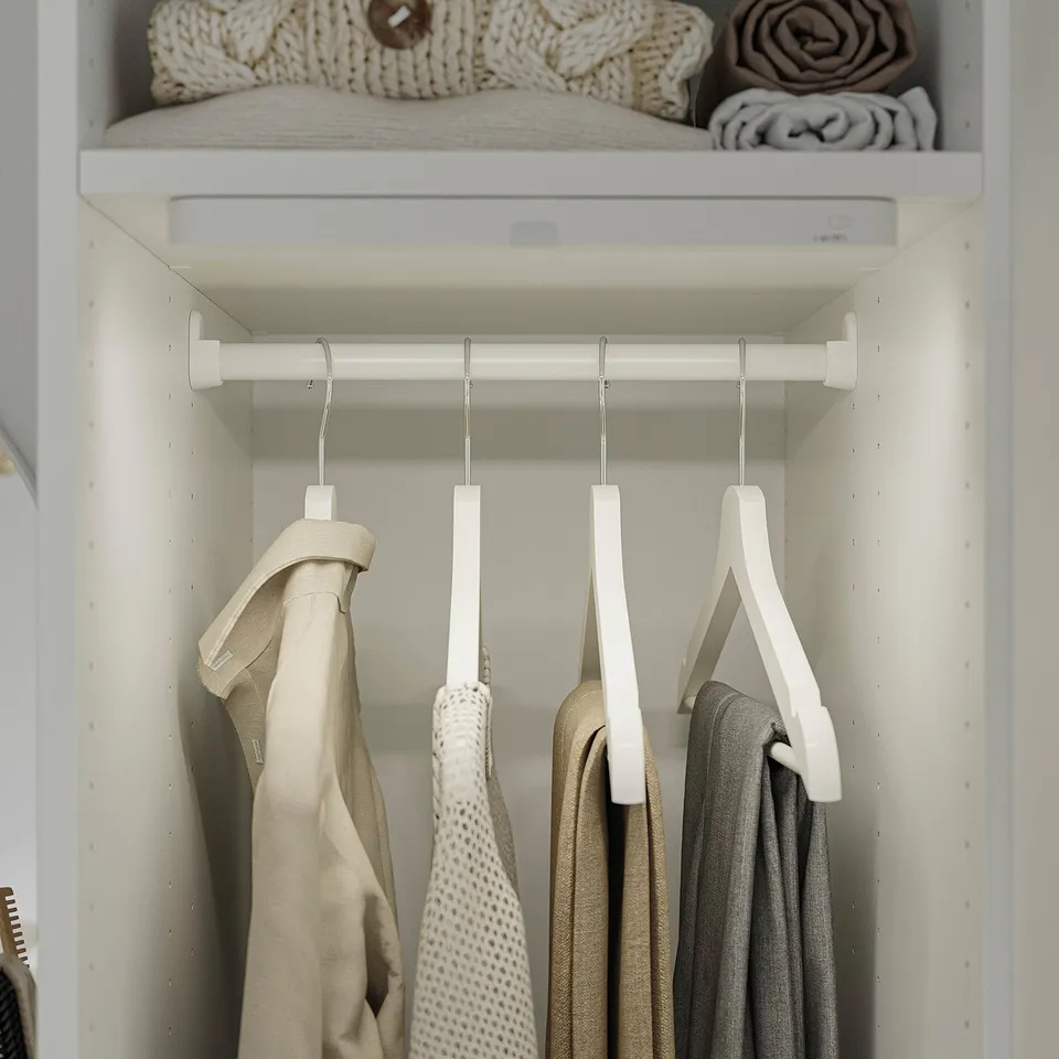 A white closet shelf with adjustable feet for floor levelling. Clothes hangers hang from a horizontal bar, holding beige and grey garments.
