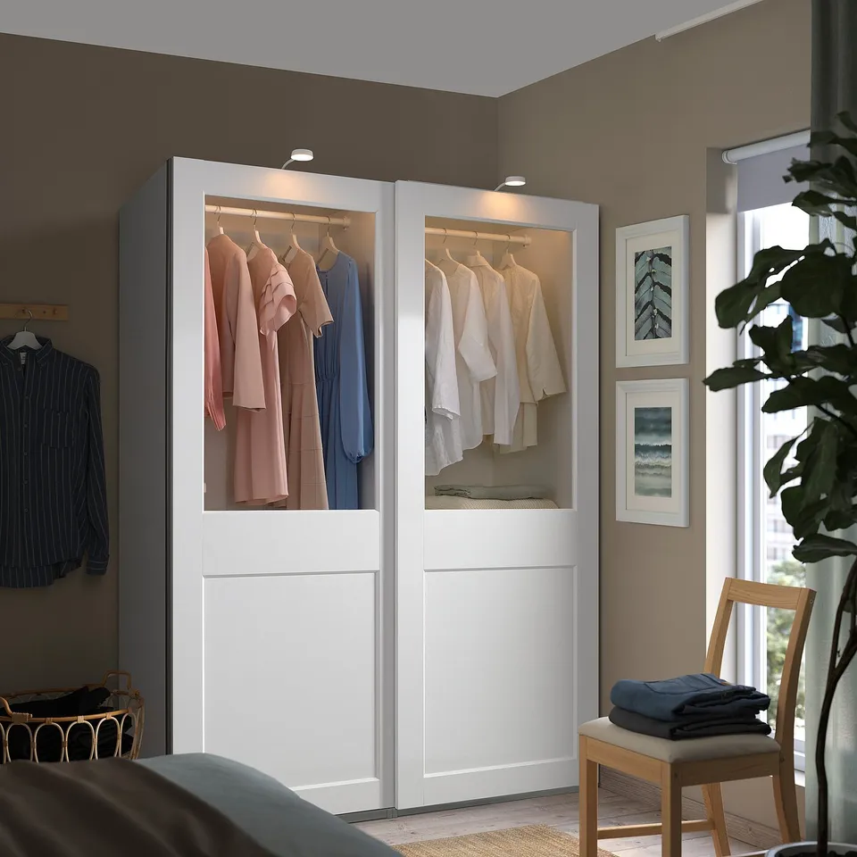 Modern white PAX wardrobe with glass doors, internal lighting, and sliding doors next to a bed.