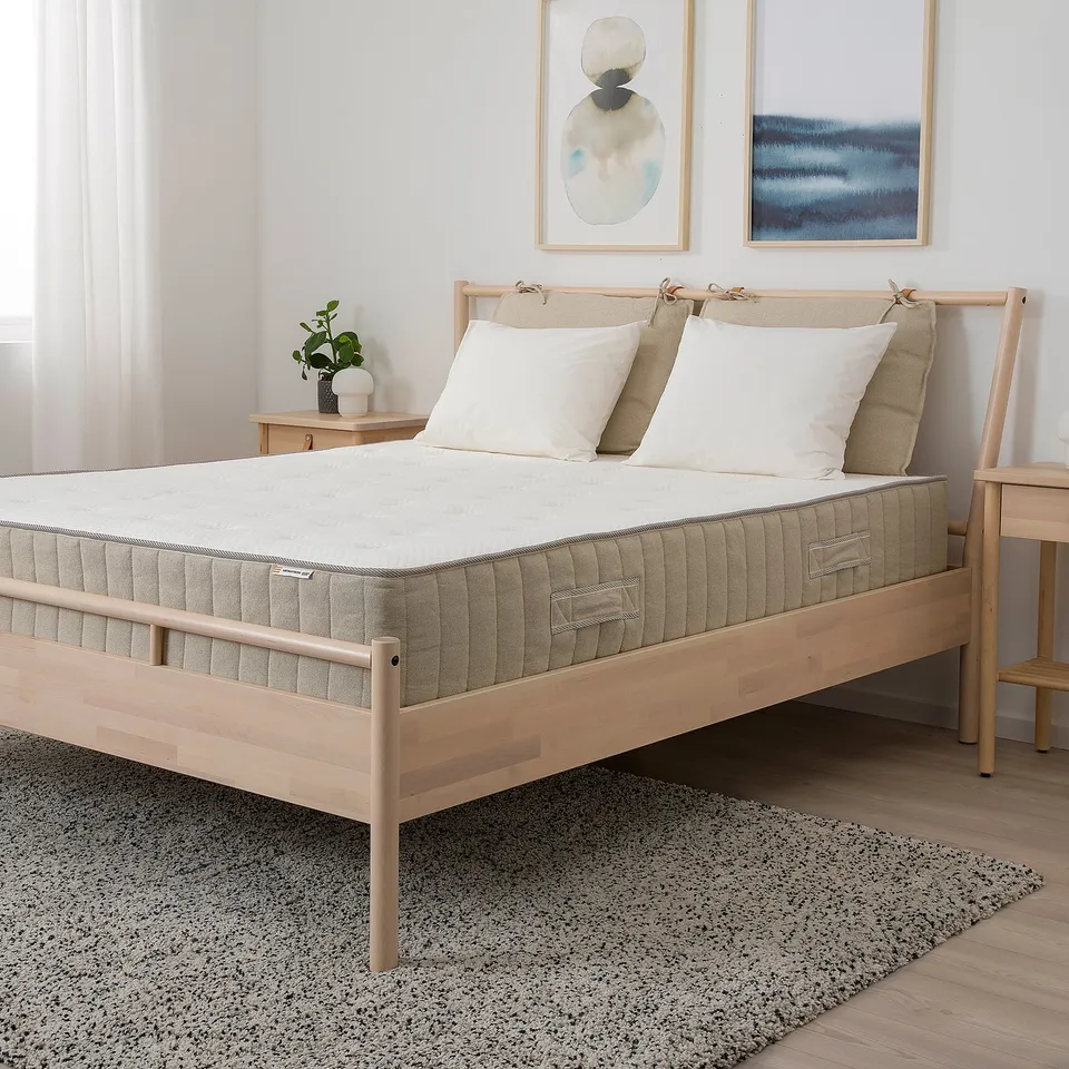 Minimalist bedroom with wooden bed, beige bedding, pillows, artwork. Left: large window. Right: bedside table with plant, lamp.