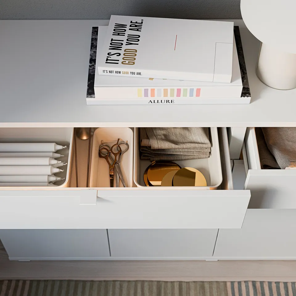 White wooden sideboard with open drawers and shelves, holding books, scissors, fabric, and golden objects.