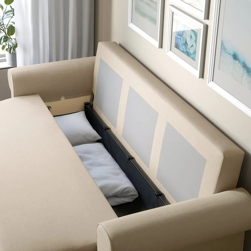 Sofa-bed with open storage compartment, revealing blanket and pillows. Light beige fabric, modern design.