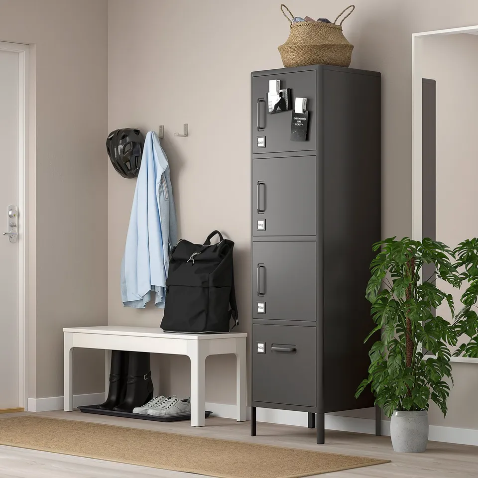 An IDÅSEN locker in grey, made of steel, with multiple compartments. It stands tall with a sleek design next to a white bench and a potted plant.