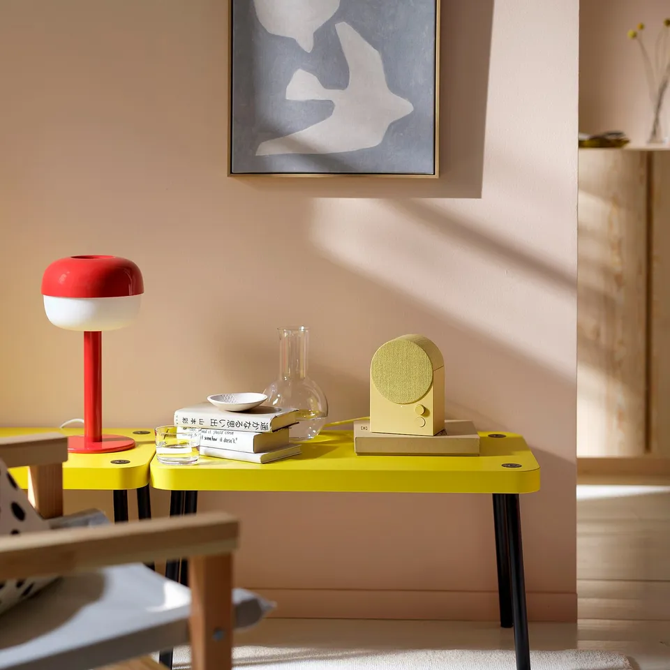 Bright yellow speaker on table with books & vases. Red lamp nearby. Art on wall. Modern, vibrant decor.