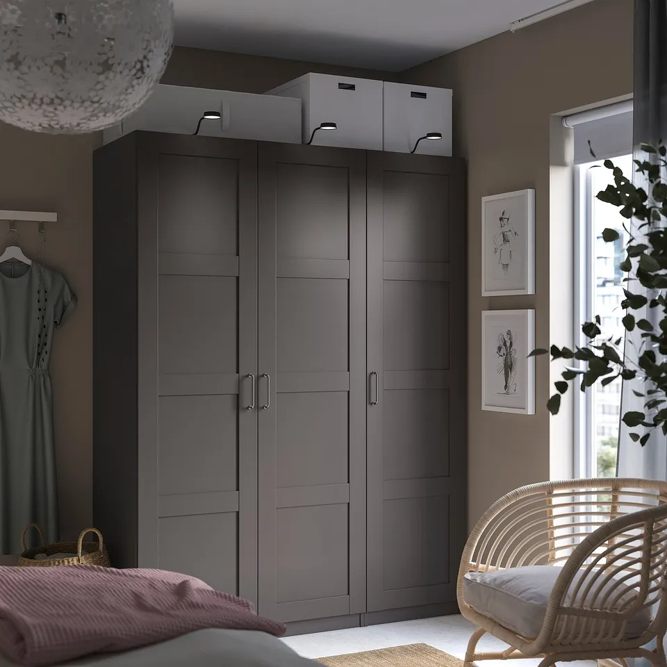 Bedroom with large dark grey BERGSBO wardrobe, white boxes, hanging clothes, wicker chair, and wall art.