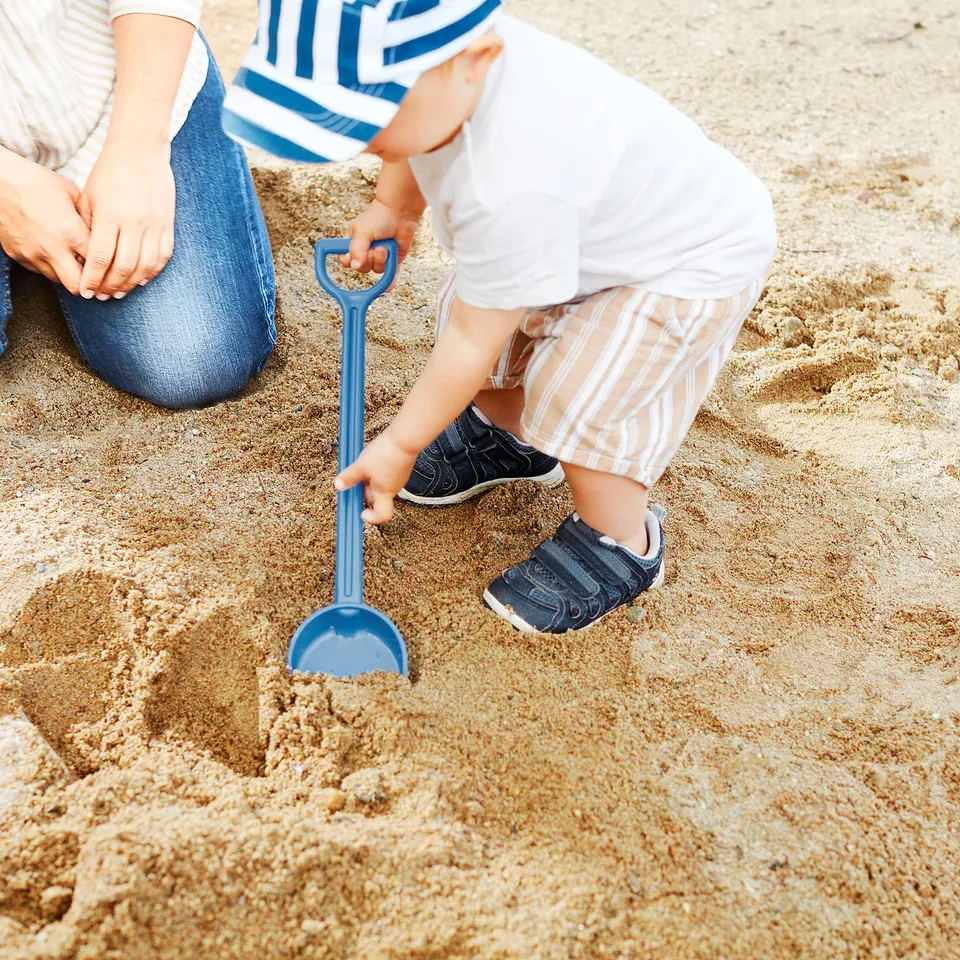 A child plays with a blue spade, digging in sandy soil, developing motor skills and creativity.