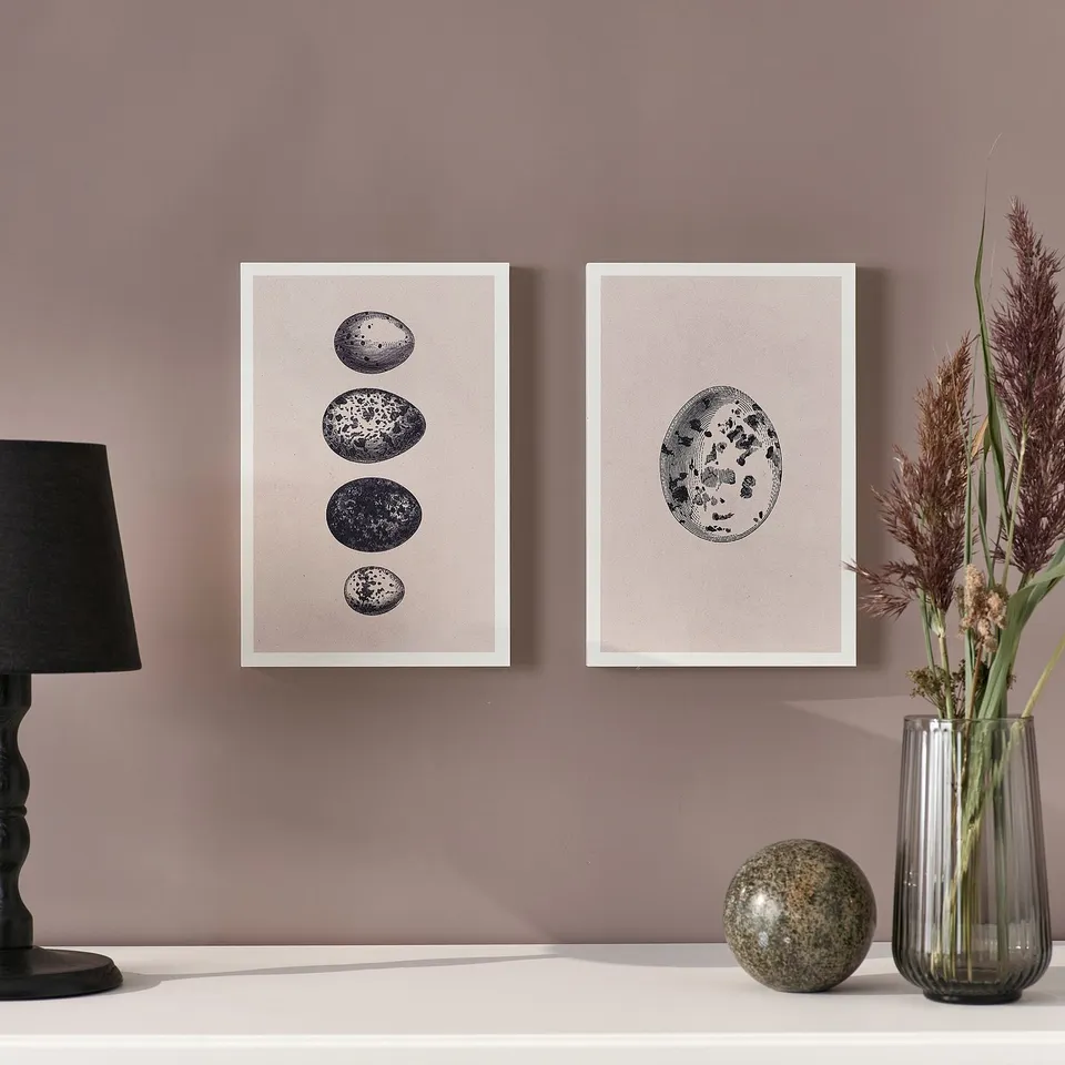 Two framed prints on wall. Left: lamp, right: vase with dried plants. Stone on table.