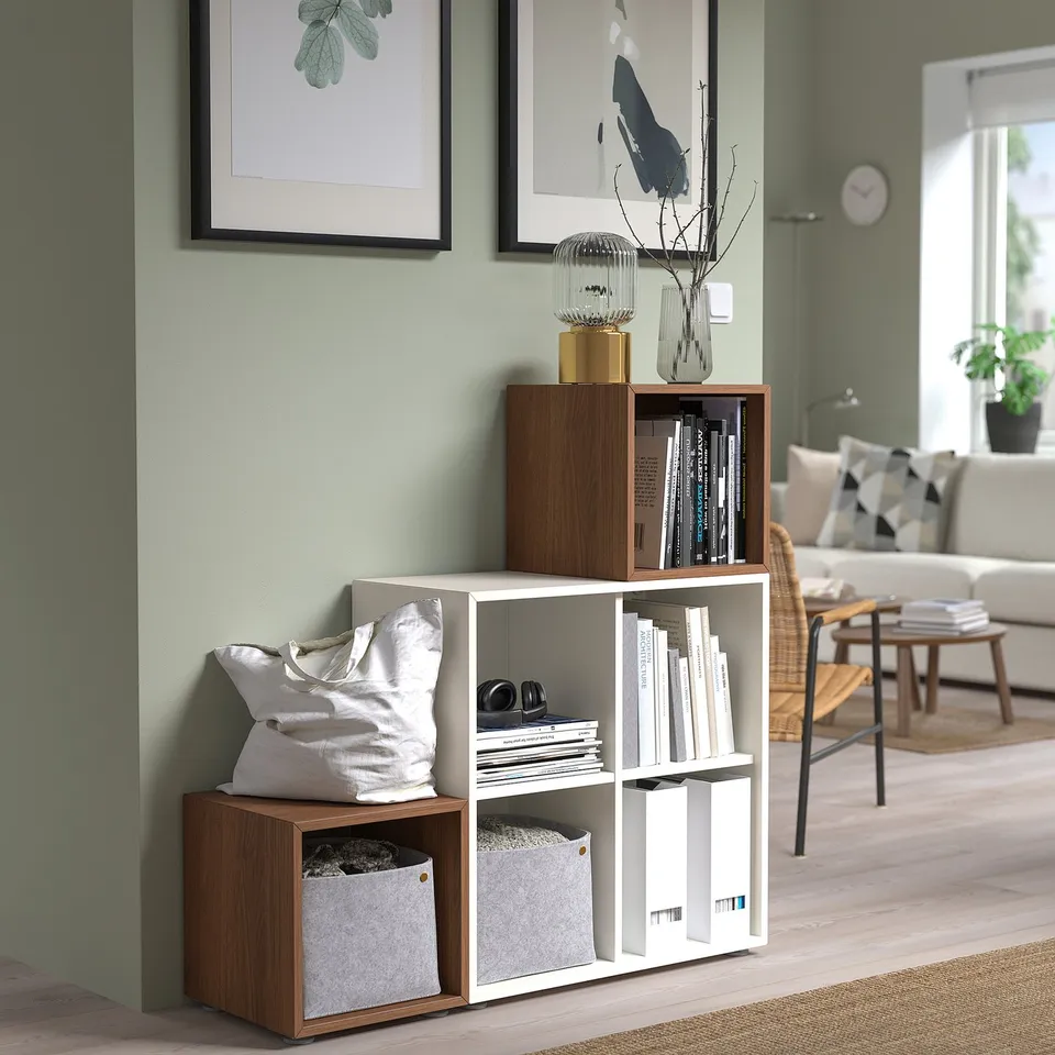 Modern living room with wood & white EKET shelves displaying books, vases, and decor; asymmetrical design suits corner.