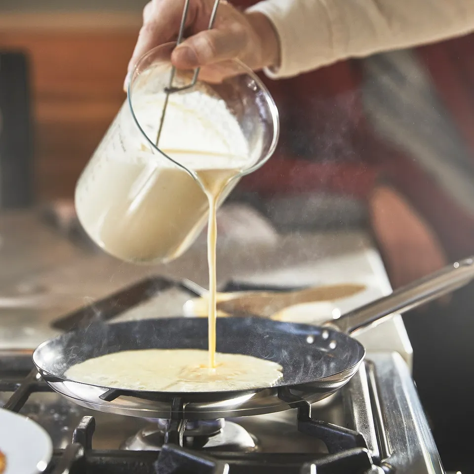 Person pouring batter from a glass jug into a frying pan on a stove, creating a crepe.